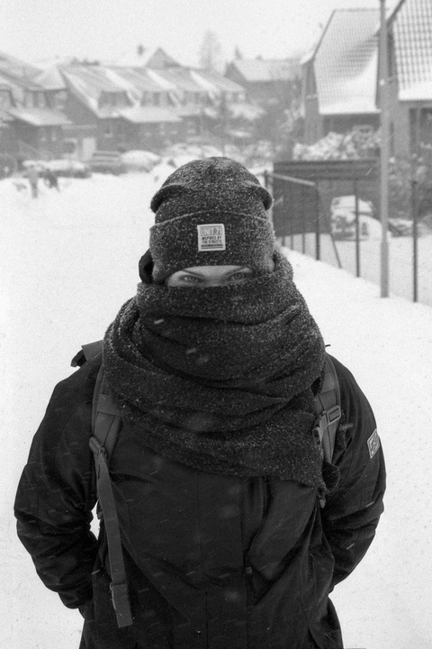 A person wearing a winter hat and scarf stands in a snowy street with houses in the background.
