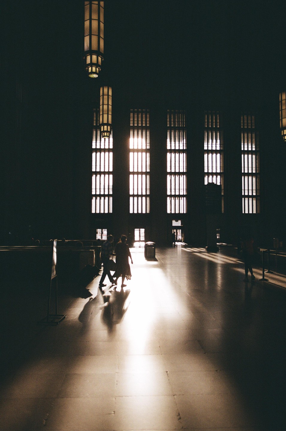 Sunlight streams through tall windows onto the floor of a large, dark hall where people walk.