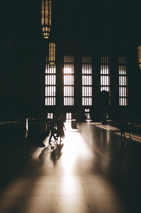 Sunlight streams through tall windows onto the floor of a large, dark hall where people walk.