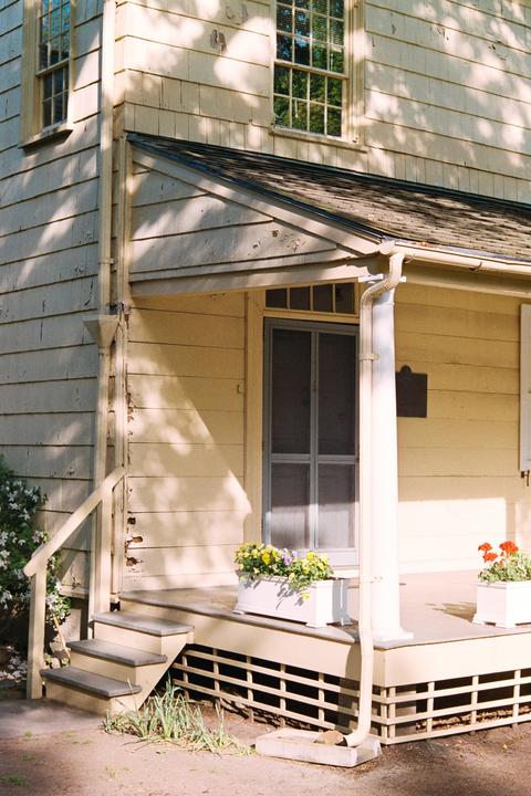 A house with peeling paint on its siding and a porch with flower boxes is visible in dappled sunlight.