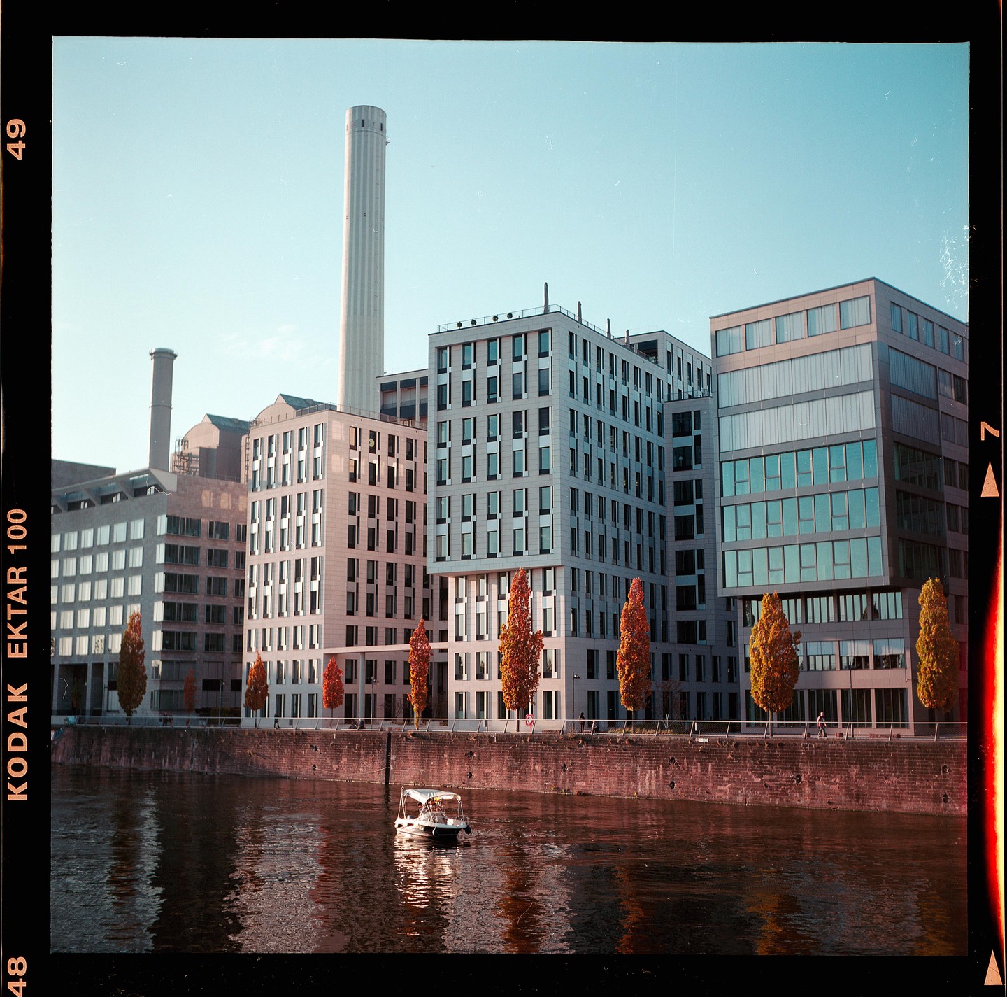A small boat floats on a river in front of modern buildings and trees with autumn foliage.