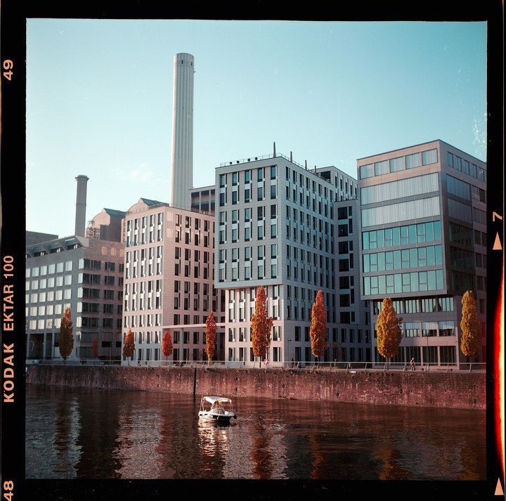 A small boat floats on a river in front of modern buildings and trees with autumn foliage.