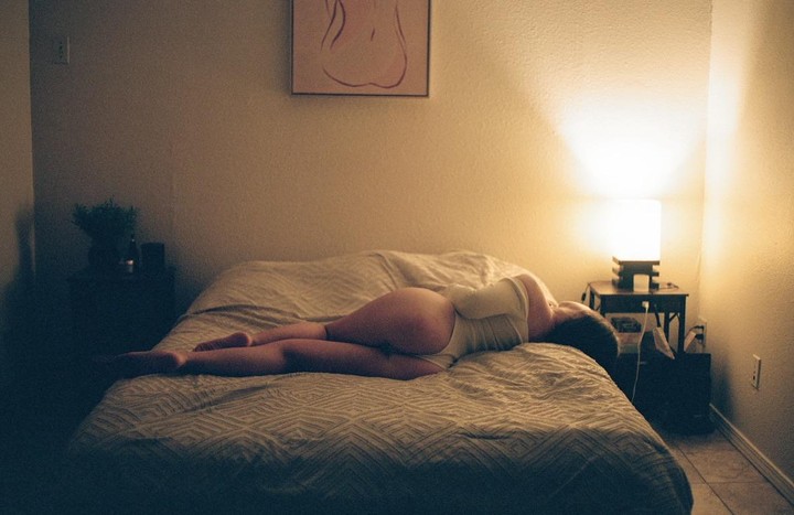 A woman lies on her side on a bed in a dimly lit room with a lamp on a nightstand.
