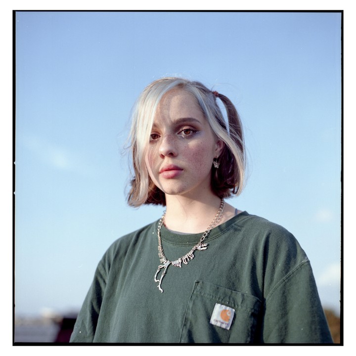 A young woman with a dinosaur necklace looks directly at the viewer under a clear sky.