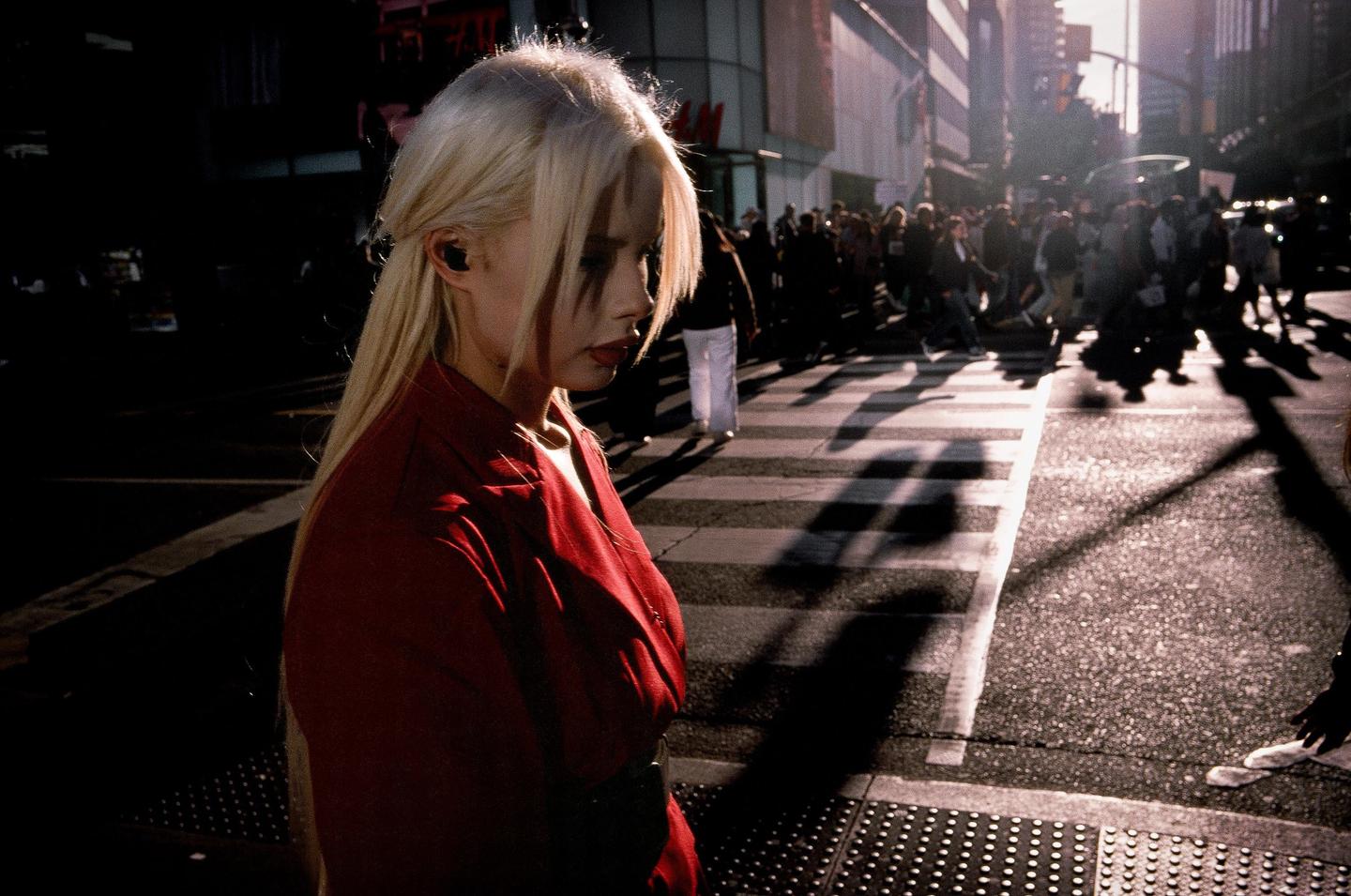 A woman with blonde hair and a red jacket stands on a city street with a crosswalk and many people in the background.