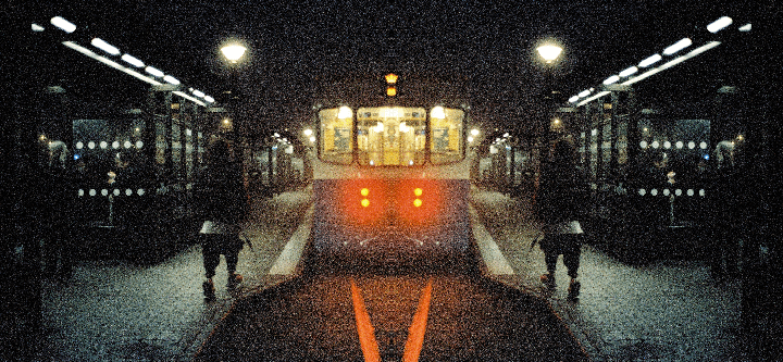 A person stands on a dark subway platform as a train approaches, its headlights glowing red.
