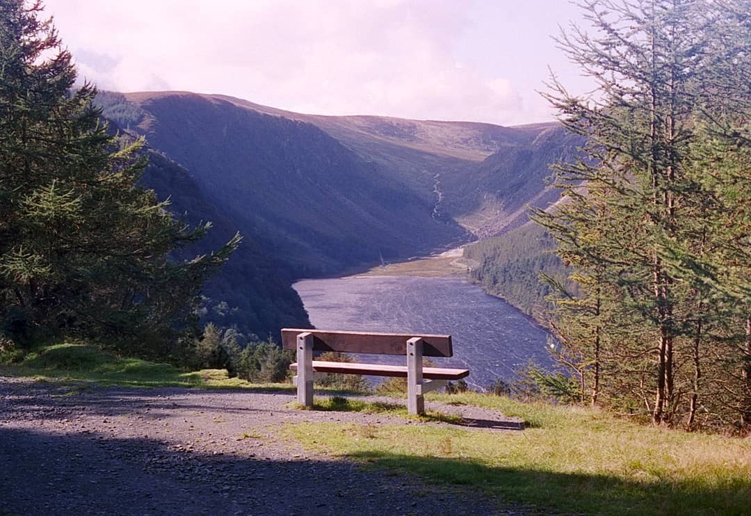 A wooden bench overlooks a valley with a lake and mountains under a hazy sky.