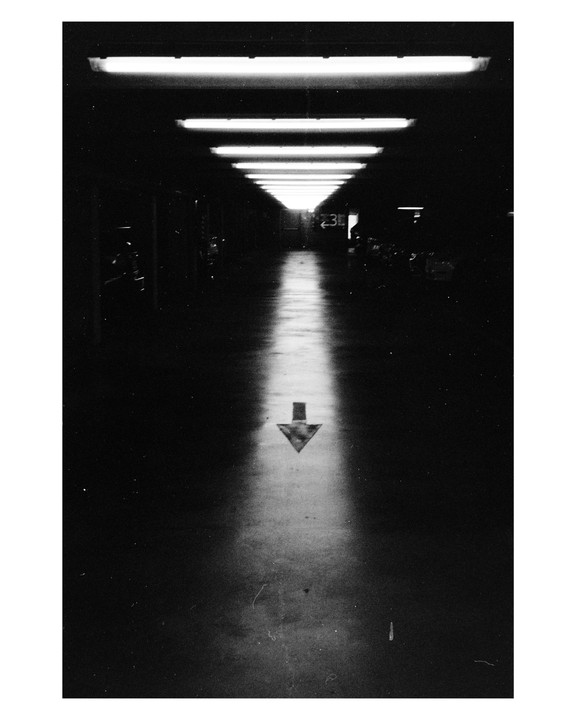 A long, dark underground parking garage is illuminated by a row of fluorescent lights on the ceiling and a bright arrow painted on the floor.