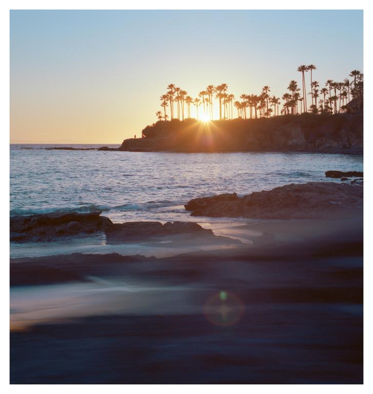 The sun sets behind a hill covered in palm trees next to a body of water with rocks in the foreground.