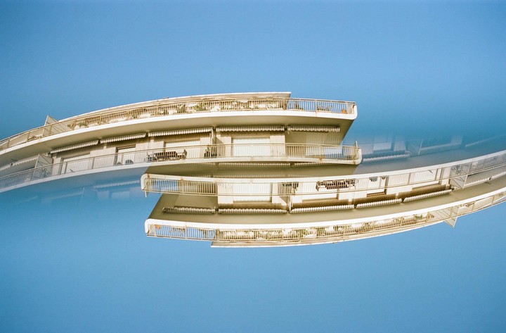 A double exposure shows a curved building with balconies against a blue sky, with the building appearing both right side up and inverted.