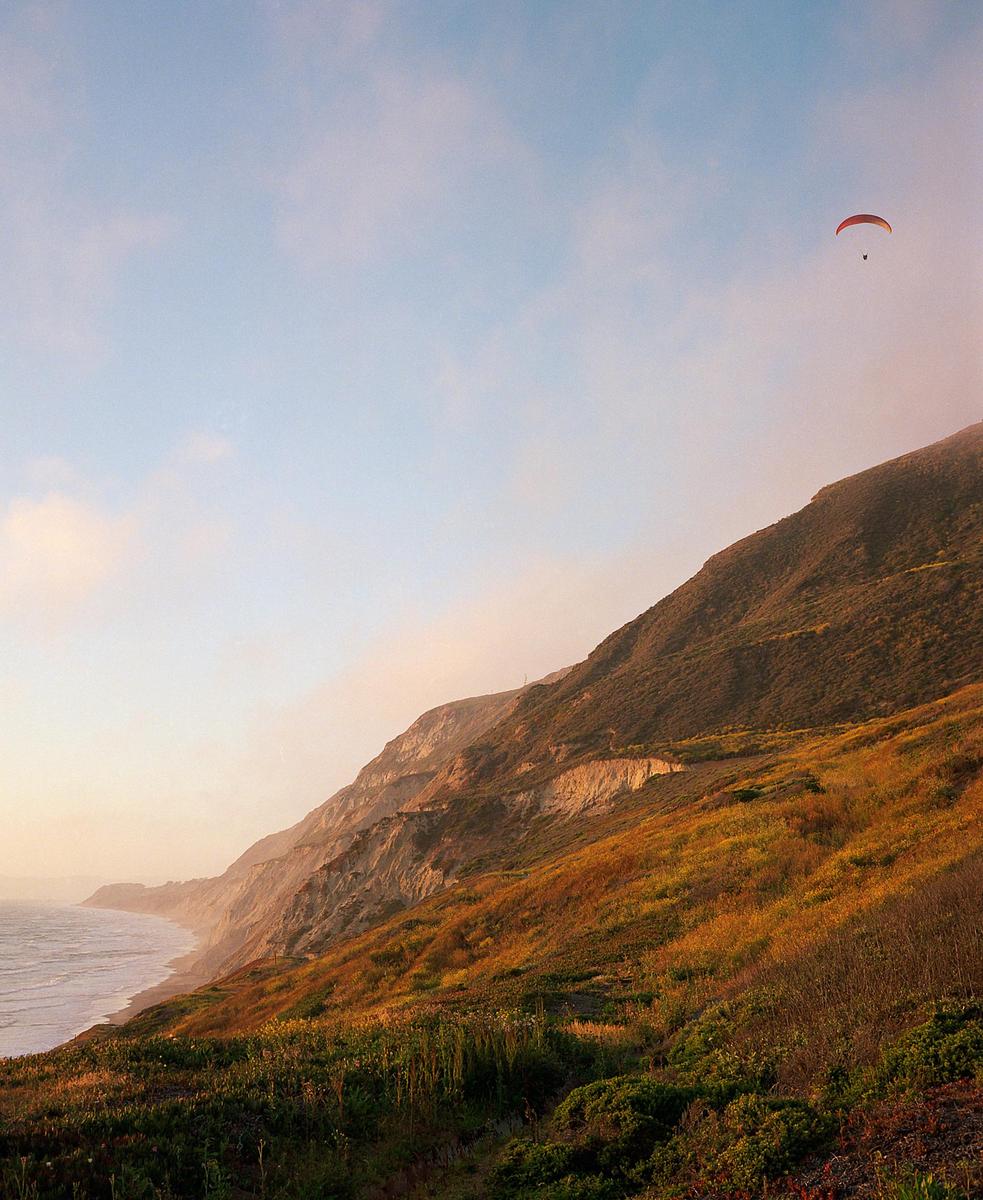 A paraglider flies over a coastal mountain landscape with the ocean visible on the left.