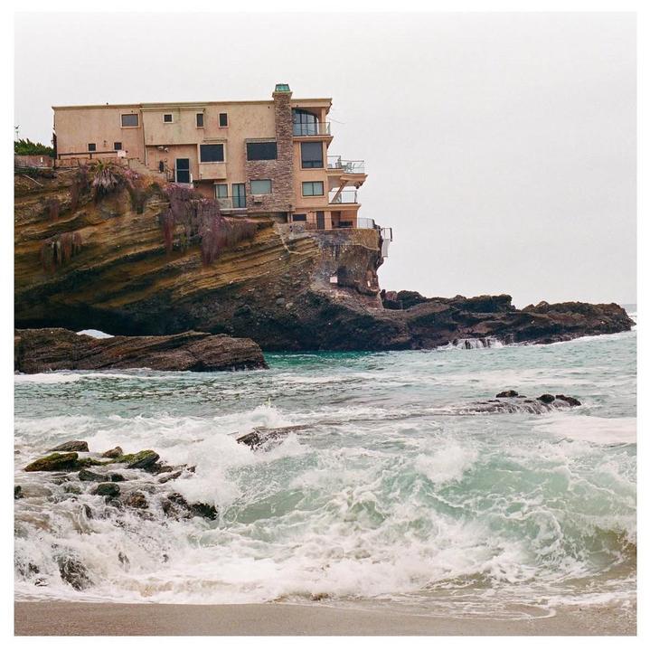 A house sits atop a rocky cliff overlooking the ocean with waves crashing on the shore.