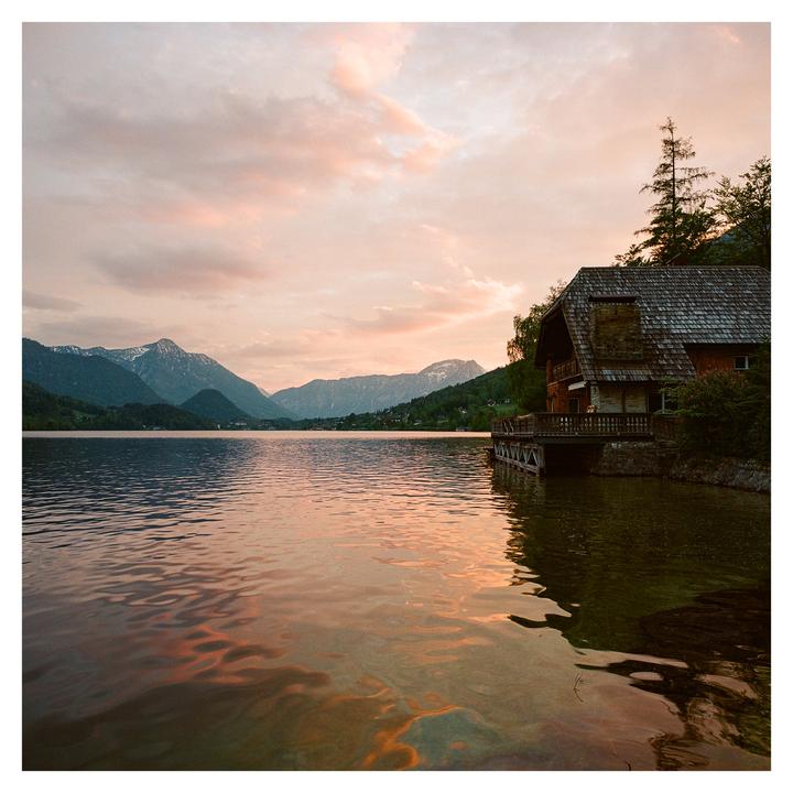 A wooden house with a dock sits on the edge of a lake with mountains in the background under a colorful sky.