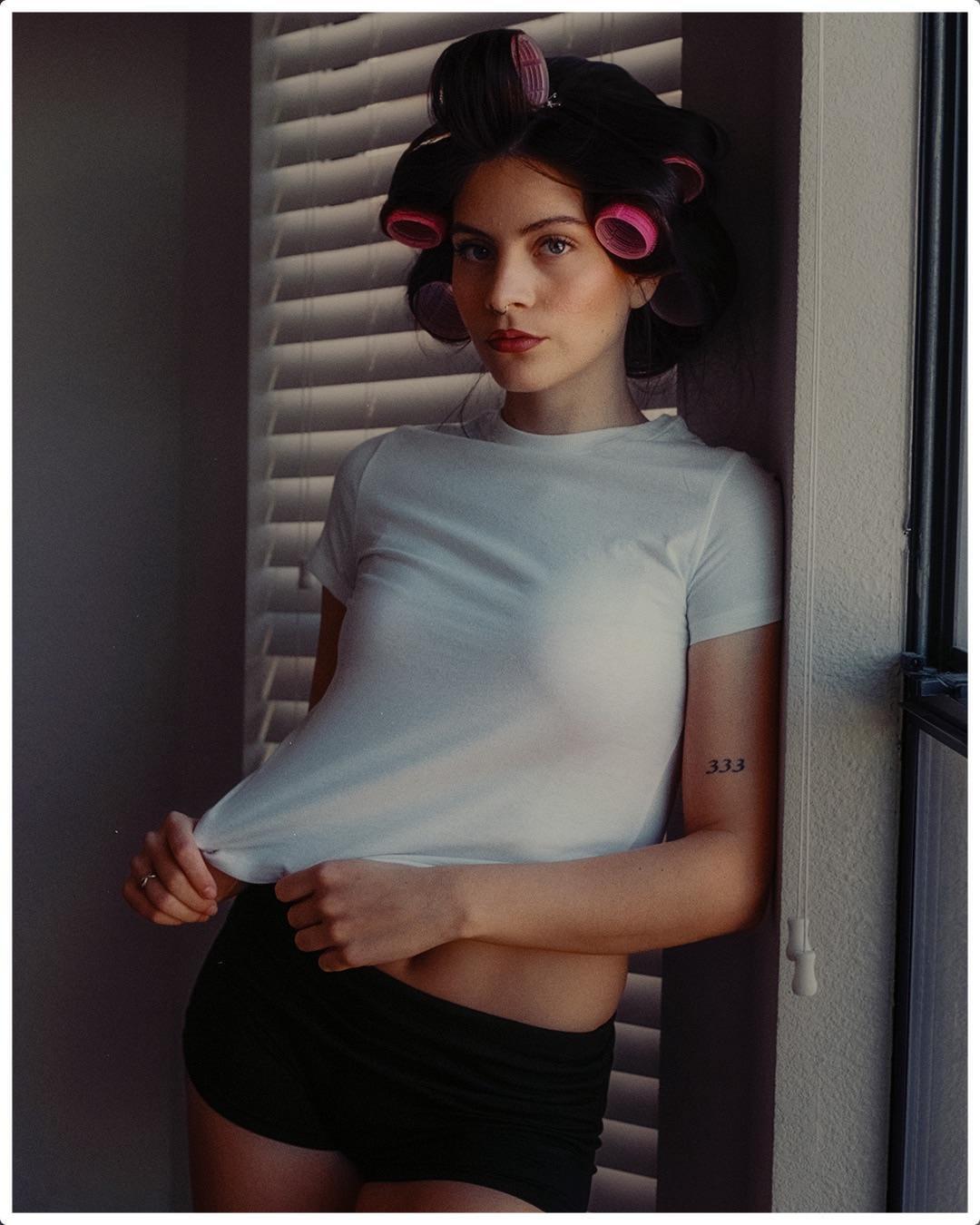 A woman with hair rollers poses in a white t-shirt and black shorts next to a window with blinds.