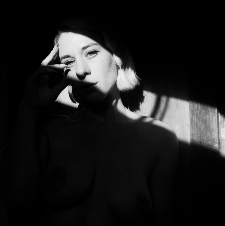 A woman with short hair poses for a self-portrait, with her hand touching her forehead, in a room with strong shadows.