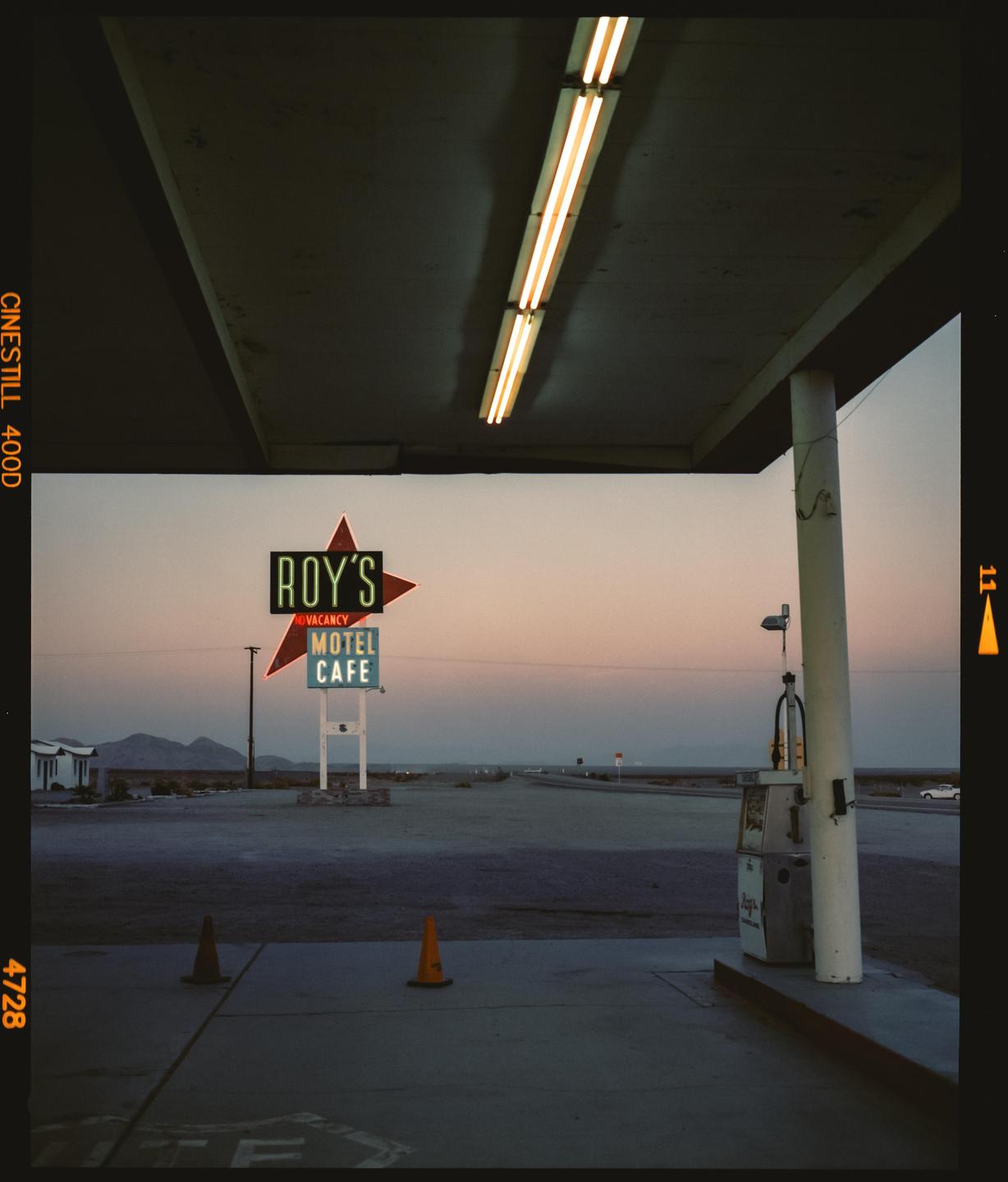 A neon sign for Roy's Motel and Cafe stands in a desert landscape at dusk, with a gas pump and traffic cones in the foreground.