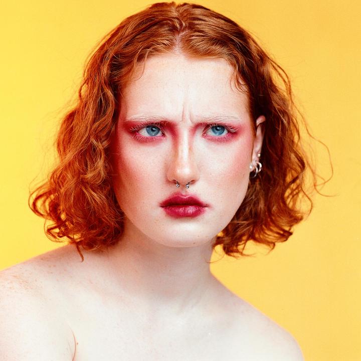 A woman with red hair and red eye makeup looks directly at the viewer with a furrowed brow against a yellow background.