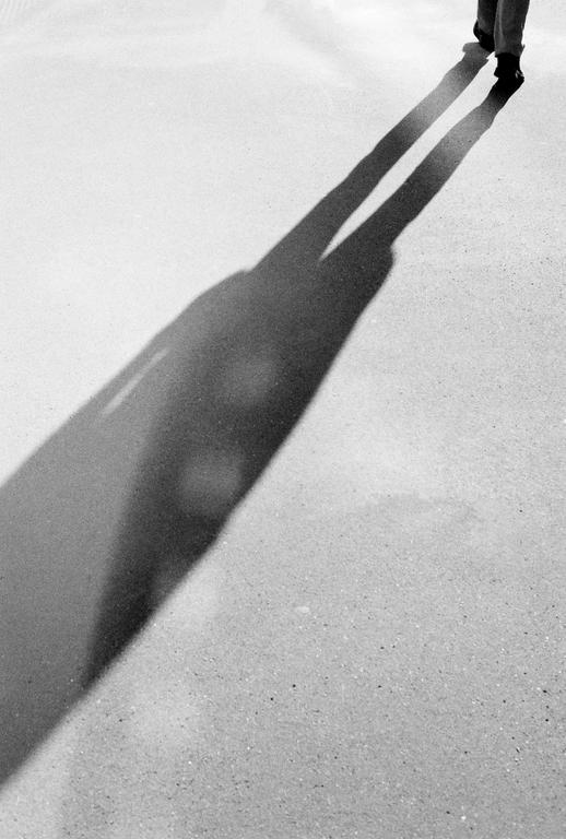 A person's long shadow stretches across a textured ground in this black and white image.