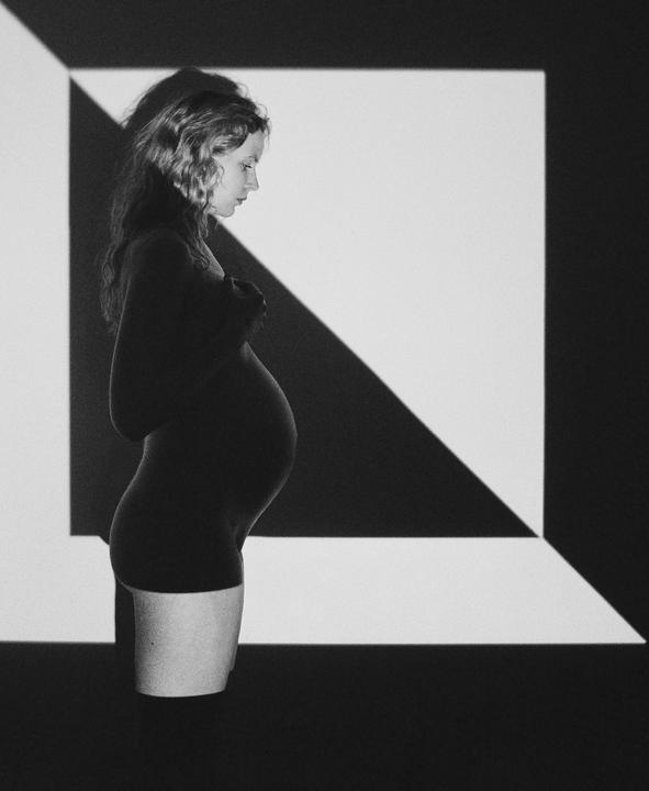 A pregnant woman stands in profile against a wall with geometric light patterns.
