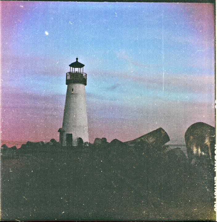 A white lighthouse stands on a rocky shore under a colorful sky.