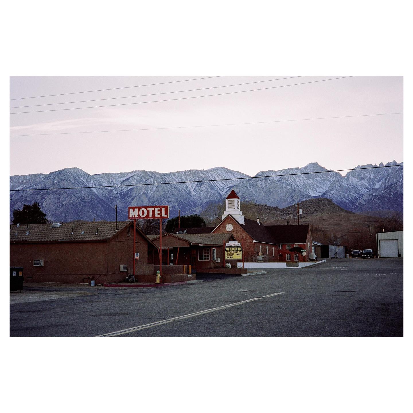 A motel sign stands in front of a brick building with a church-like steeple, with snow-capped mountains in the background.