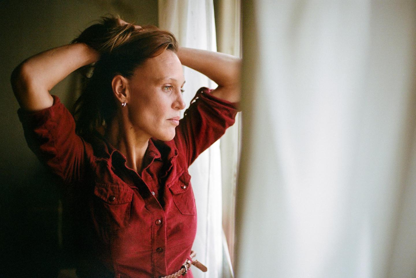 A woman in a red shirt looks out a window, with her hands in her hair.