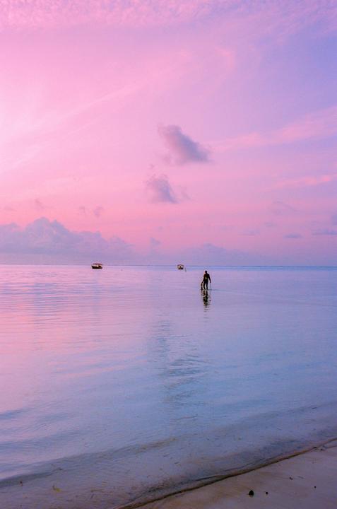 A man stands in shallow water with two boats in the distance under a vibrant pink and purple sky.