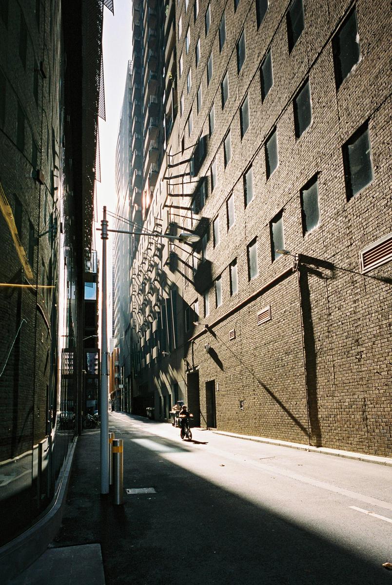 A person rides a bicycle down a narrow alley between tall brick buildings, with sunlight creating strong shadows on the right wall.