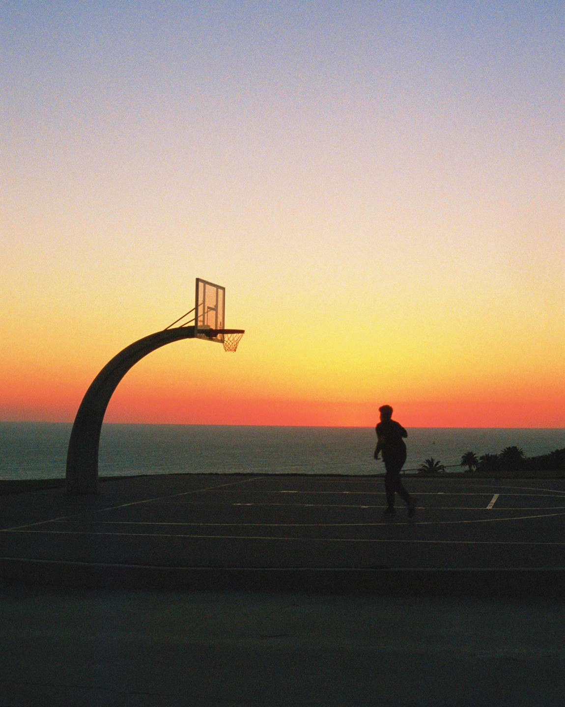 A man stands on a basketball court overlooking the ocean at sunset.