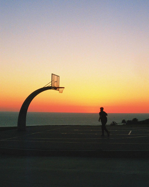 A man stands on a basketball court overlooking the ocean at sunset.