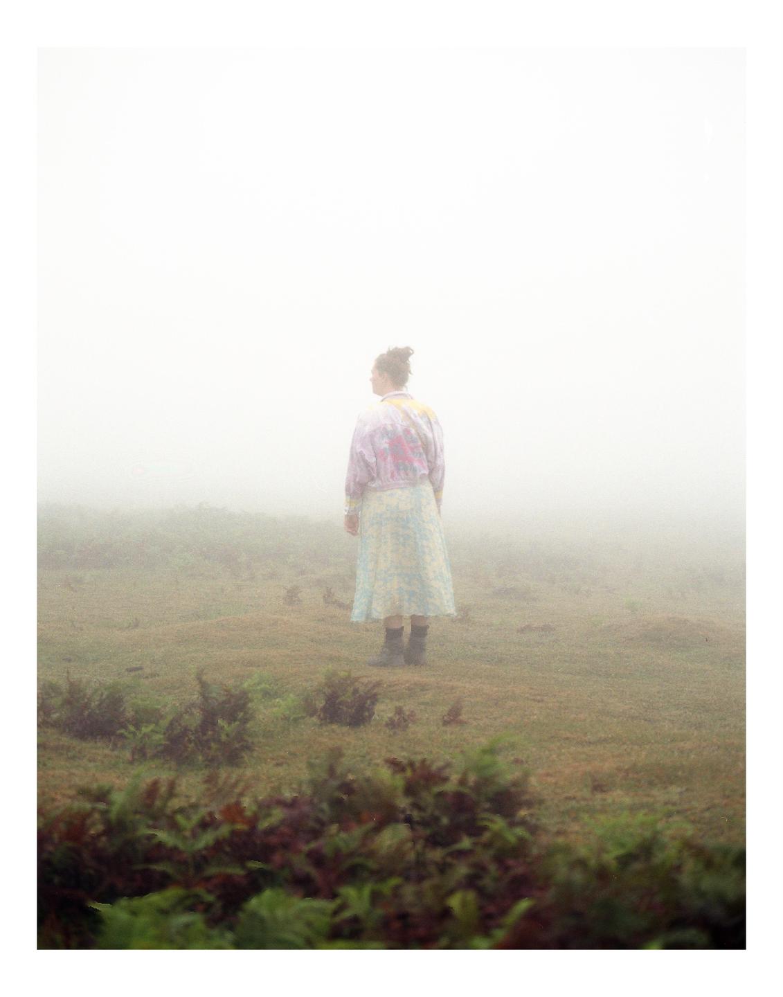 A woman stands in a field of low-lying plants, looking out into a thick fog.