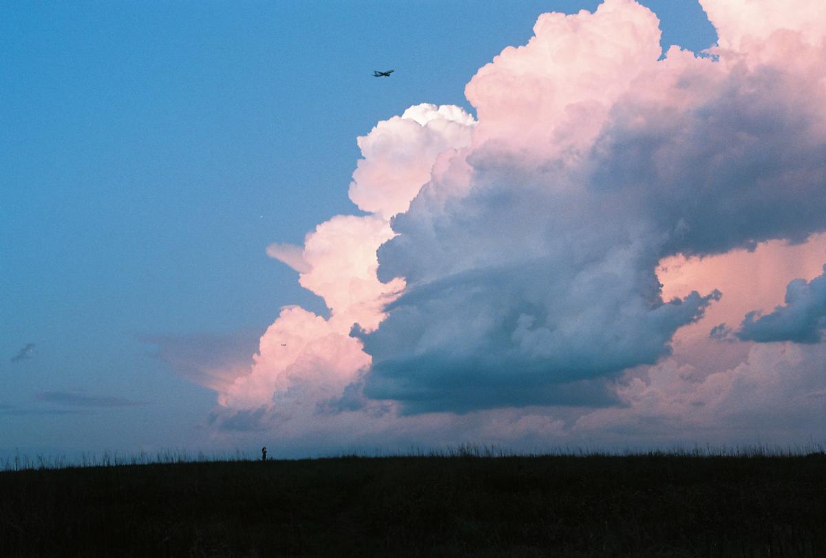A small airplane flies above a field with a person standing in the distance under a sky filled with large, colorful clouds.