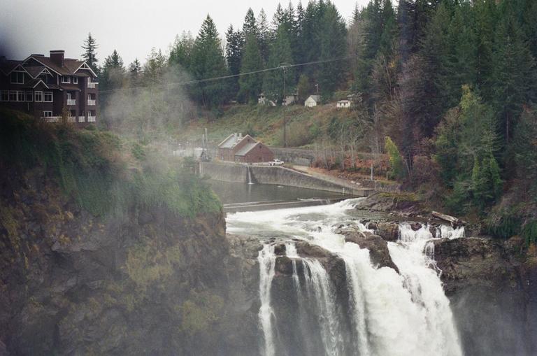 A large waterfall cascades over rocks with mist rising, and buildings are visible on the forested cliffs above.