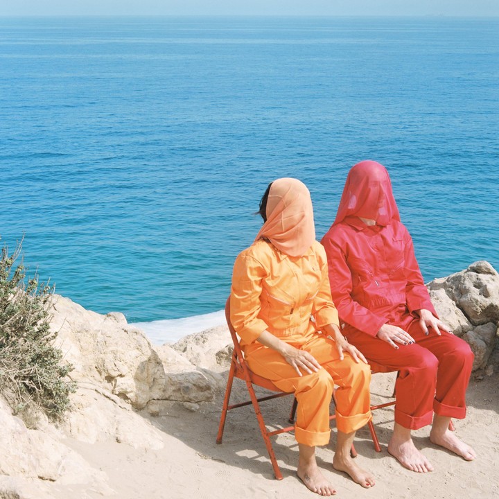 Two people in colorful jumpsuits sit on chairs facing the ocean, one with a peach-colored veil and the other with a red veil.