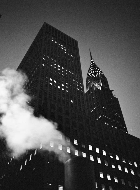 Steam rises in front of a tall building with many illuminated windows, with the Chrysler Building visible in the background.
