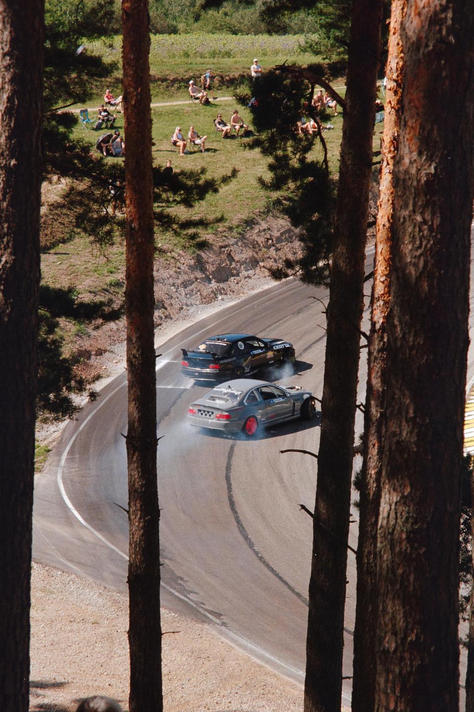 Two cars drift around a curve on a road with spectators watching from a grassy hill.