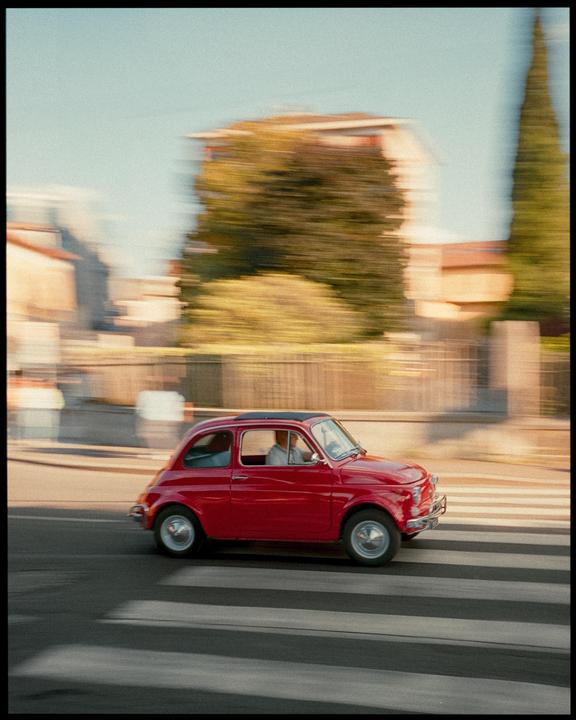 A red car drives across a crosswalk with blurred buildings and trees in the background.
