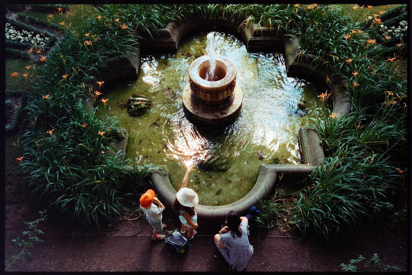 Three people are gathered around a stone fountain with green water and orange flowers in a garden.