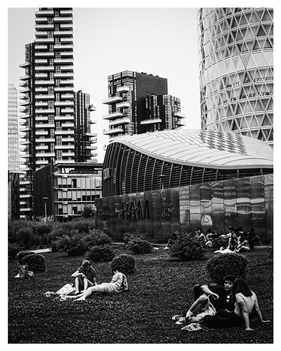 People relax on a grassy area surrounded by modern buildings in Milan.