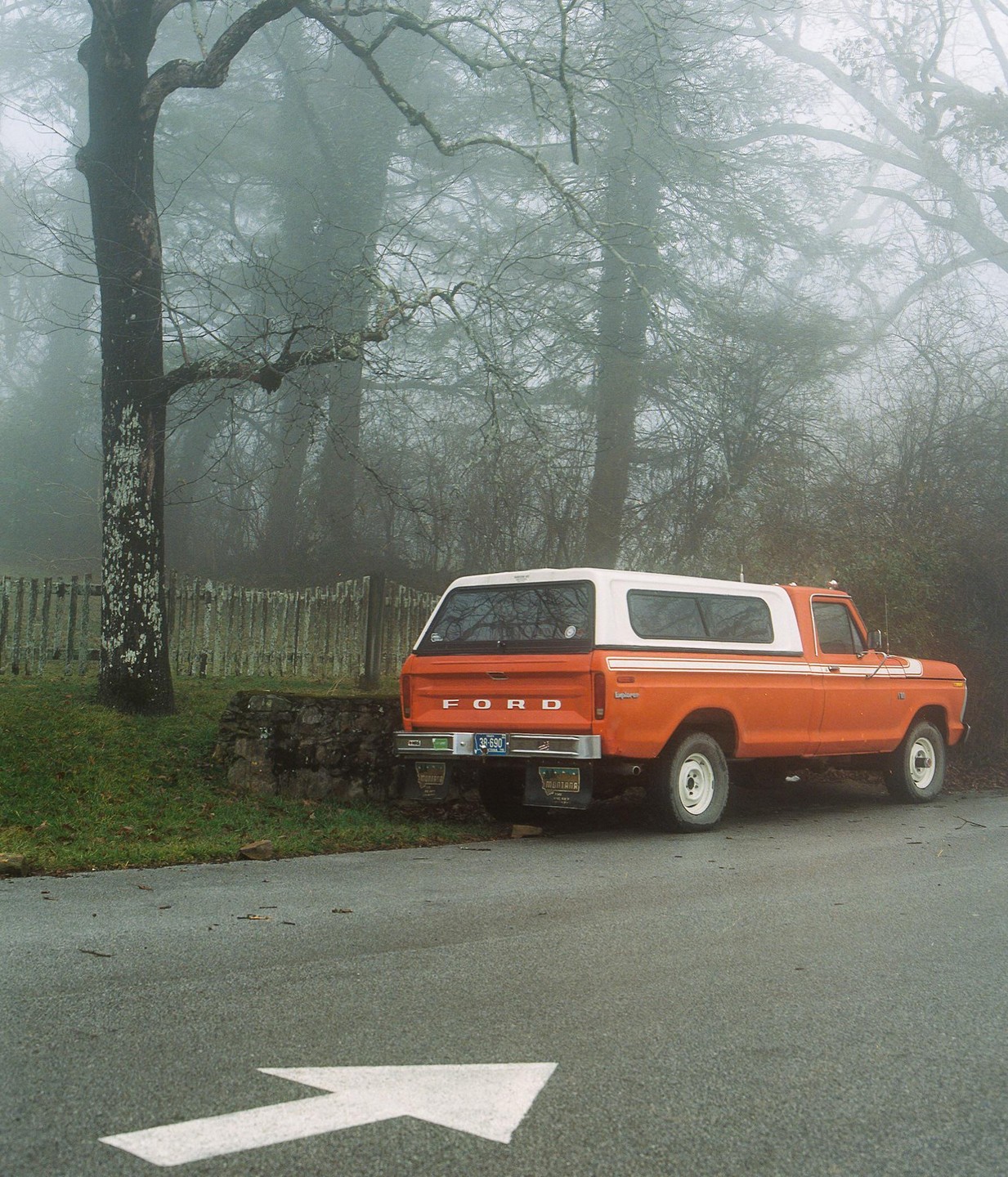An orange and white pickup truck is parked on the side of a road with a white arrow painted on the asphalt, surrounded by trees and fog.