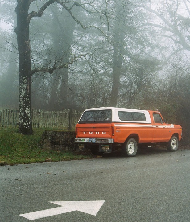 An orange and white pickup truck is parked on the side of a road with a white arrow painted on the asphalt, surrounded by trees and fog.