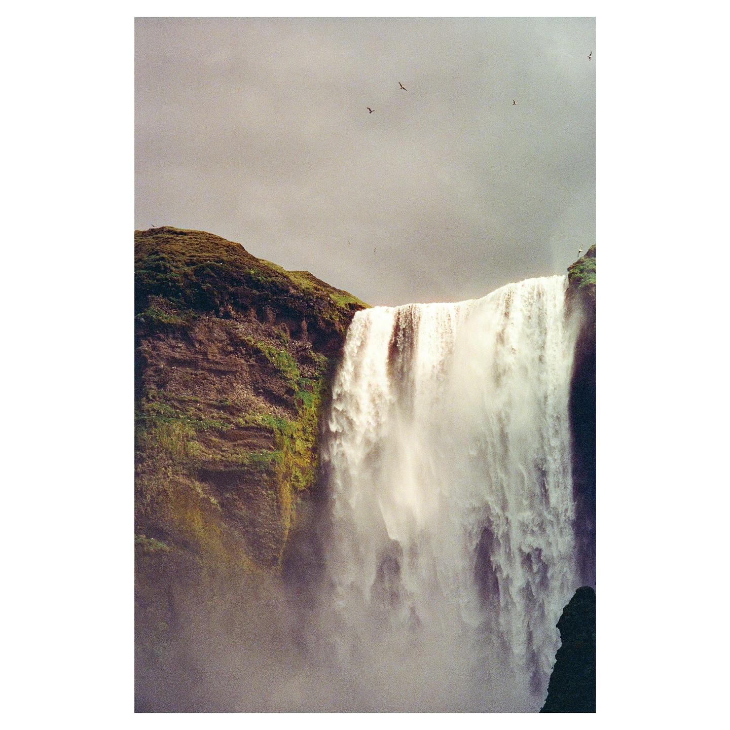 A powerful waterfall cascades down a mossy cliff under a cloudy sky with birds flying overhead.
