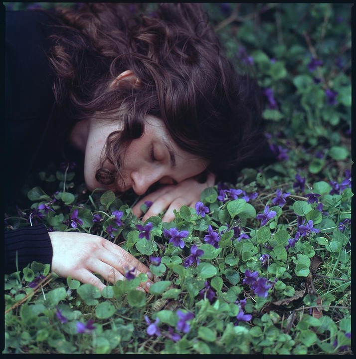 A woman with closed eyes is lying in a field of purple flowers and green leaves.