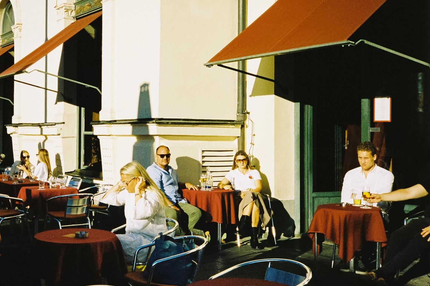 People sit at tables with red tablecloths under awnings on a sunny street.
