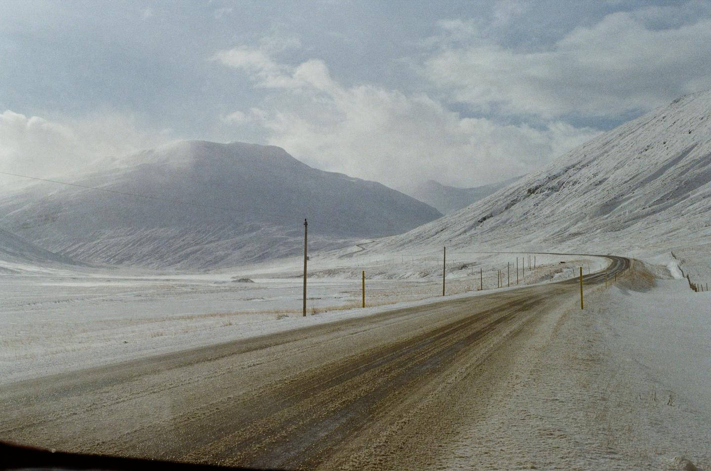 A winding road cuts through a snow-covered valley with mountains under a cloudy sky.