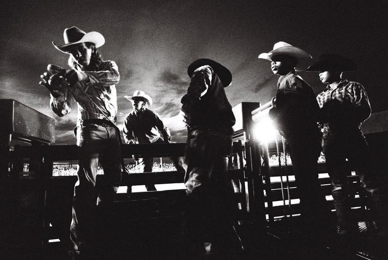Several men in cowboy hats are standing on a wooden structure at night, with one man in the foreground adjusting something in his hands.