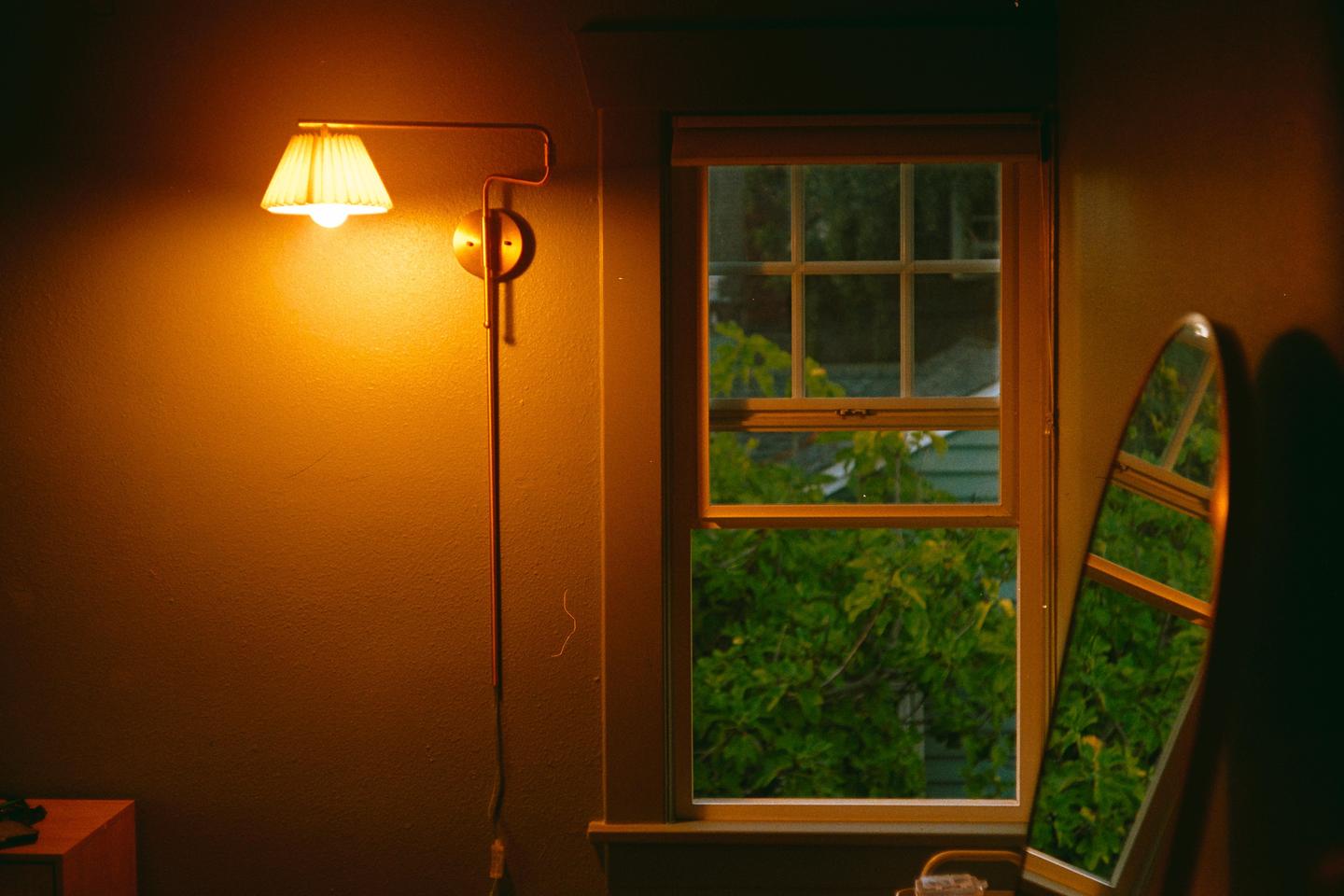 A wall sconce illuminates a room with a window looking out onto green foliage, next to a mirror reflecting the window.