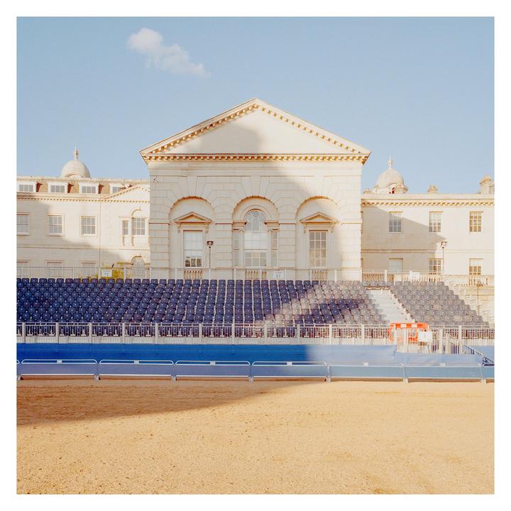 A large, ornate building with a triangular pediment stands behind rows of empty blue stadium seats.
