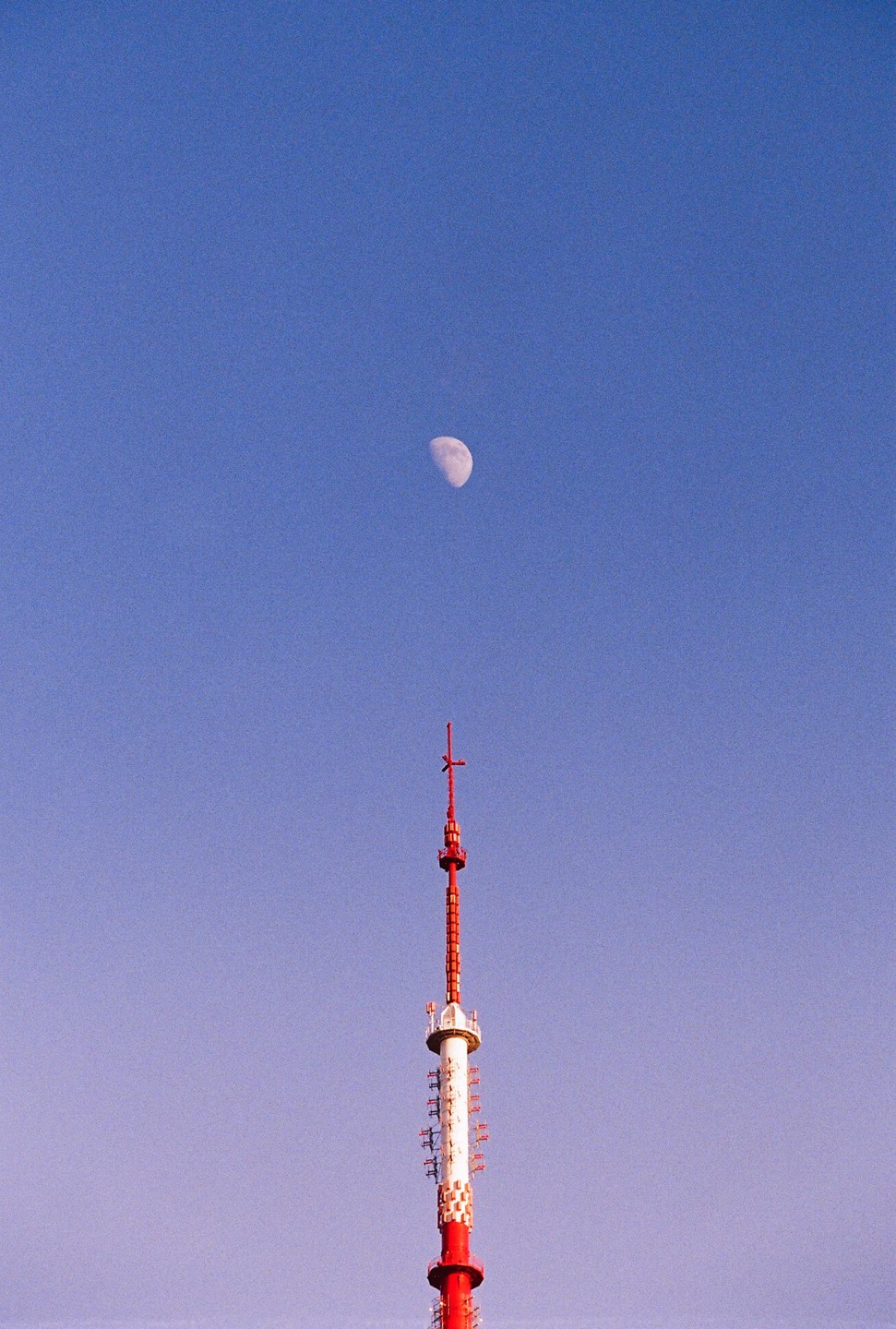 A red and white communication tower stands tall against a clear blue sky with a crescent moon visible above it.
