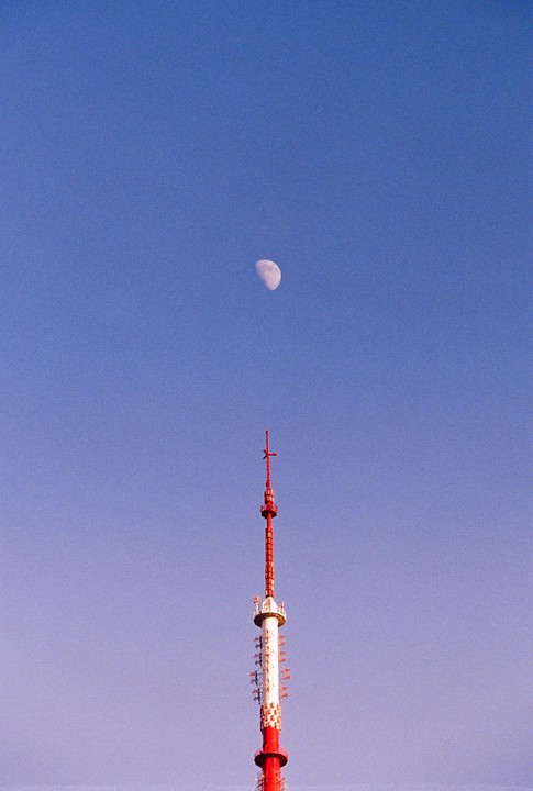 A red and white communication tower stands tall against a clear blue sky with a crescent moon visible above it.